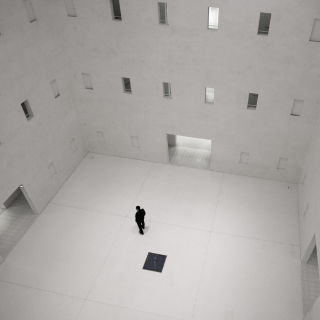 Lone man stands in a high-ceilinged white room with many windows, looking at the wall, in black and white